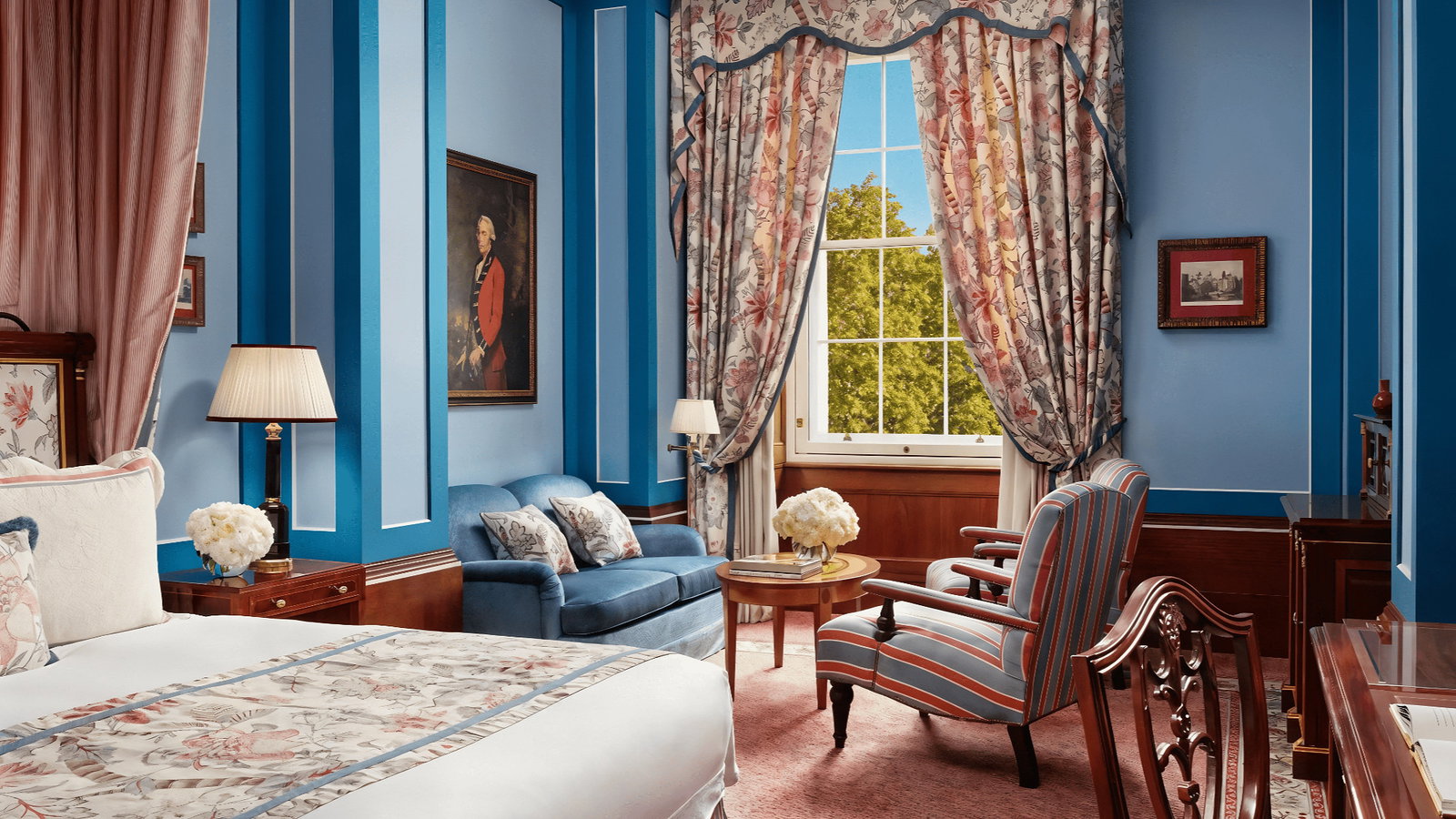 Elegant bedroom with blue walls and classic decor. Floral curtains frame a sunny window. Cozy bed, blue sofa, and red-striped chair.