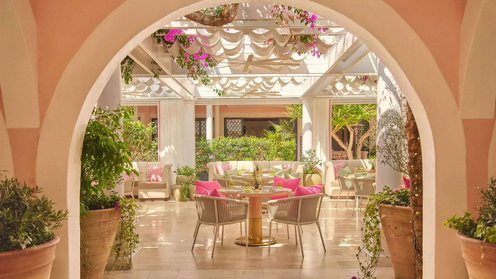 Bright, airy patio with pink accents, wicker chairs, and lush greenery. Arched entryway frames tables and couches under a sunlit canopy.