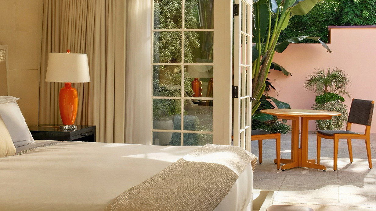 Elegant bedroom with white bed and orange lamp. Open glass doors lead to a patio with green plants, a wooden table, and chairs. Cozy ambiance.