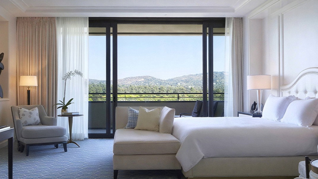 Elegant hotel room with a large bed, armchair, and a view of green hills through floor-to-ceiling windows. Soft blue and beige tones. Calm mood.