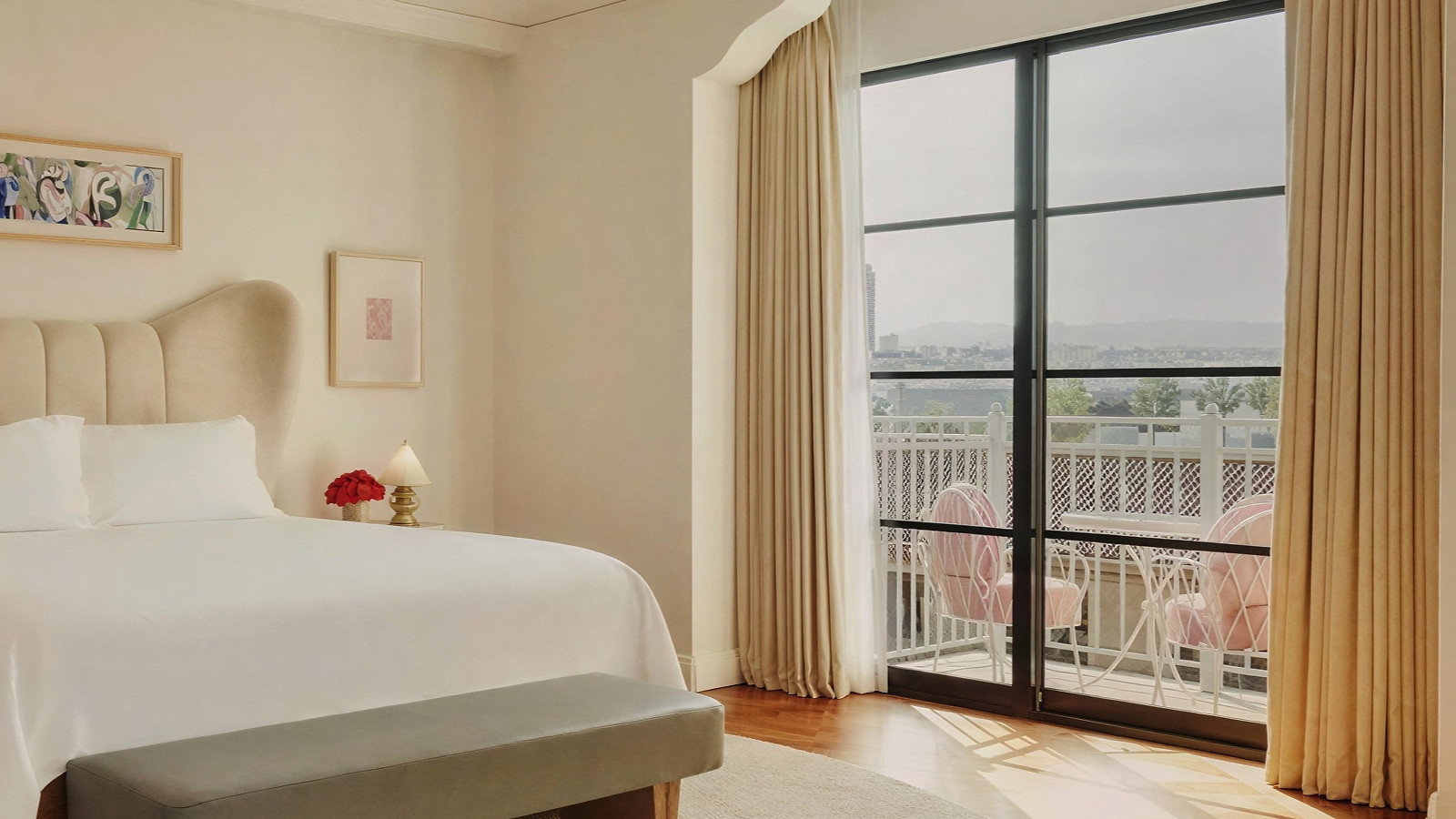 Elegant bedroom with a cream bed, soft lighting, and a window view of a patio with pink chairs. Beige curtains and light wood flooring.