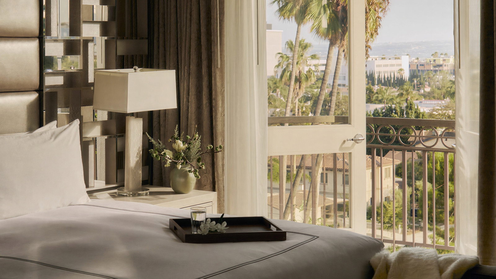 Luxurious hotel room with a made bed, decorative pillows, and a tray. Open balcony door reveals palm trees and a sunny outdoor view.