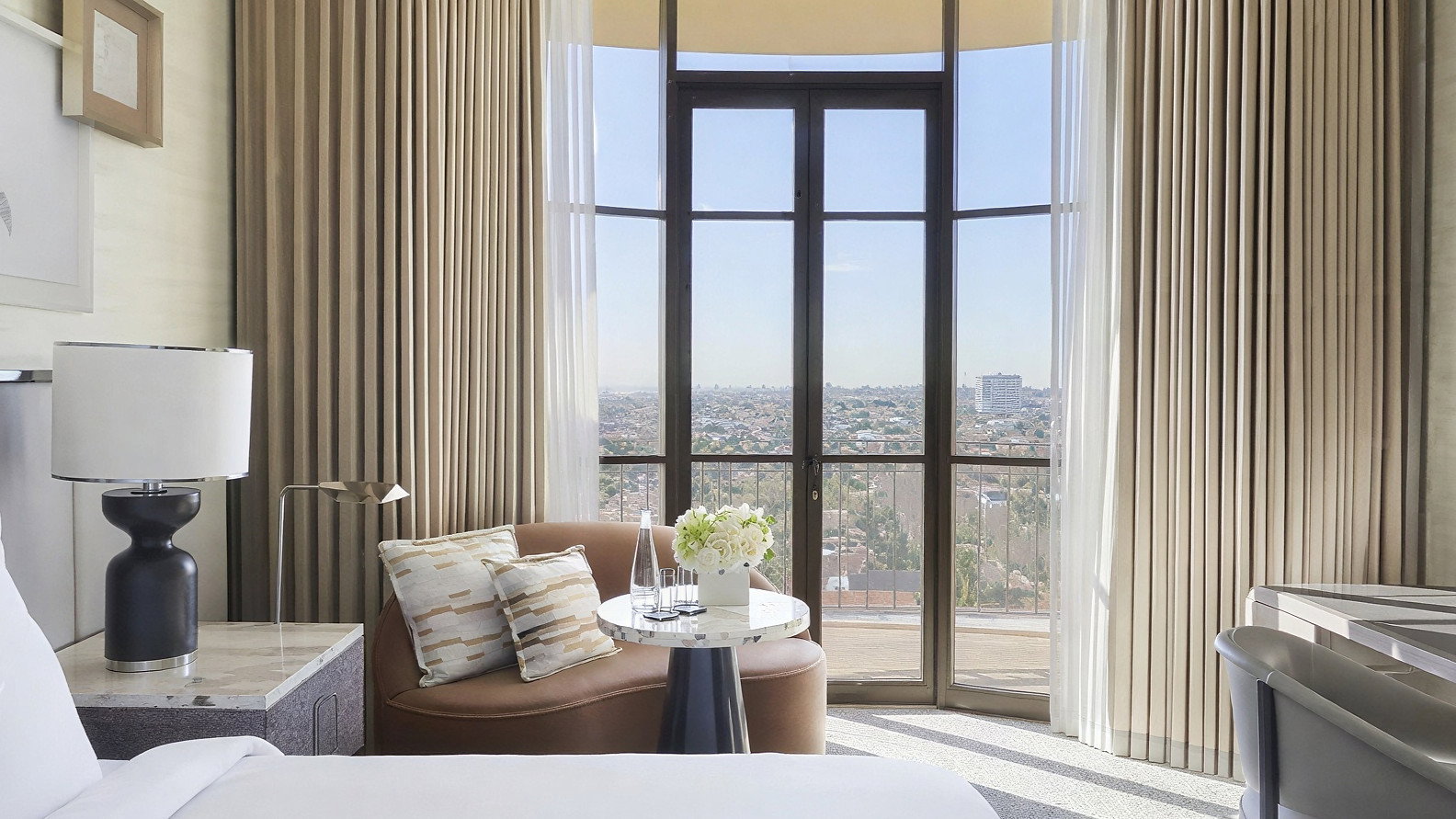Elegant hotel room with a large window view of a cityscape. Beige curtains, cozy armchair, white bed, and light decor create a calm mood.