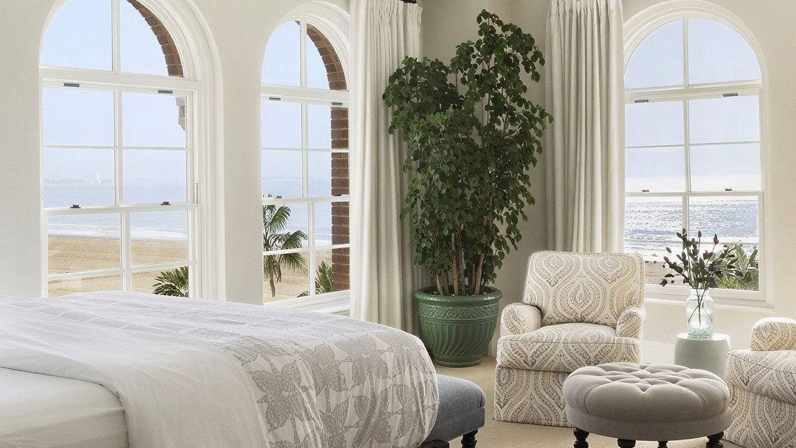 Elegant bedroom with ocean view, featuring a bed with patterned linens, tufted ottoman, armchairs, large plant, and cream curtains.