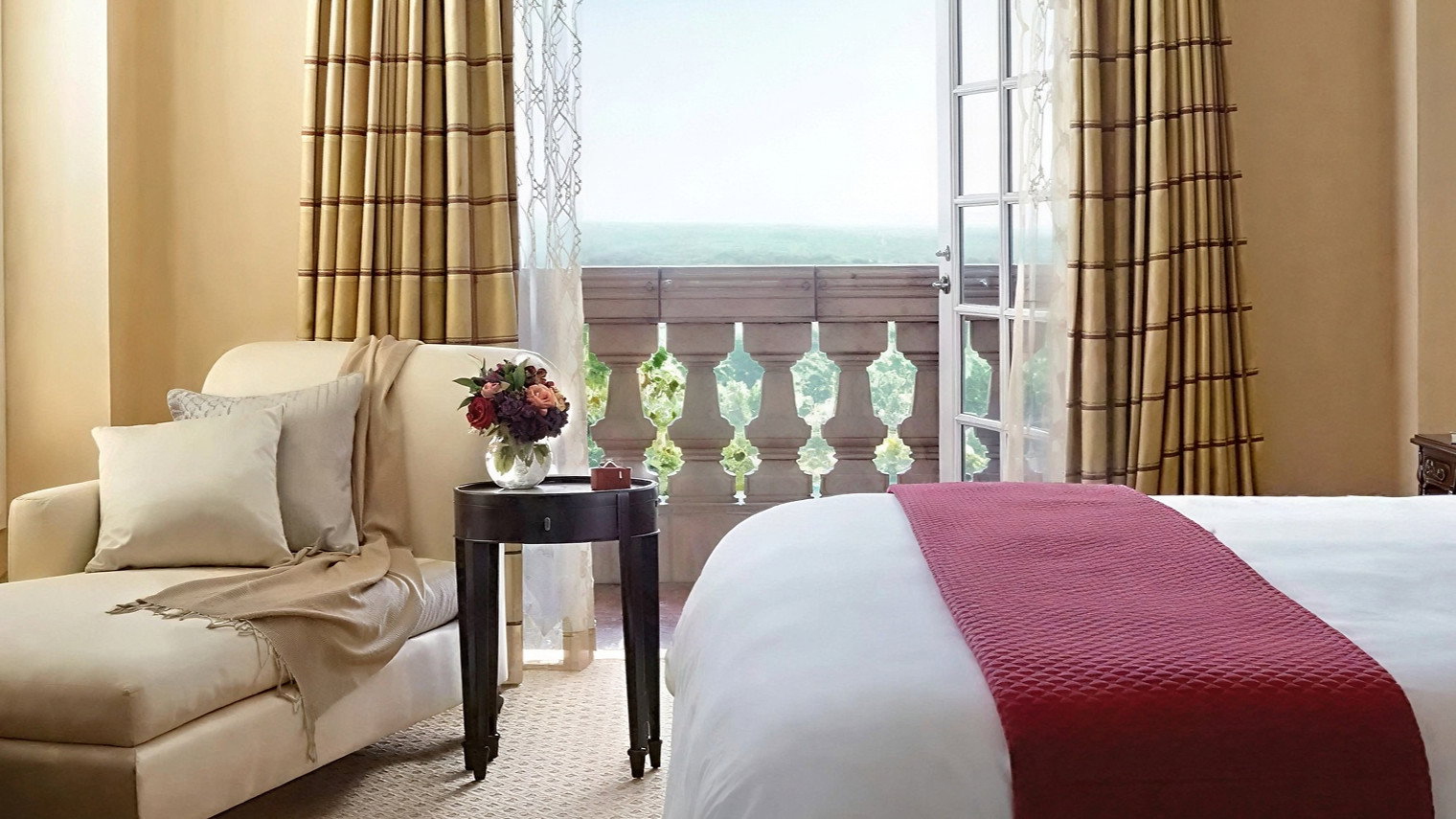 Luxurious hotel room with beige chaise, red accented bed, and open doors to balcony overlooking greenery. Elegant ambiance with warm tones.