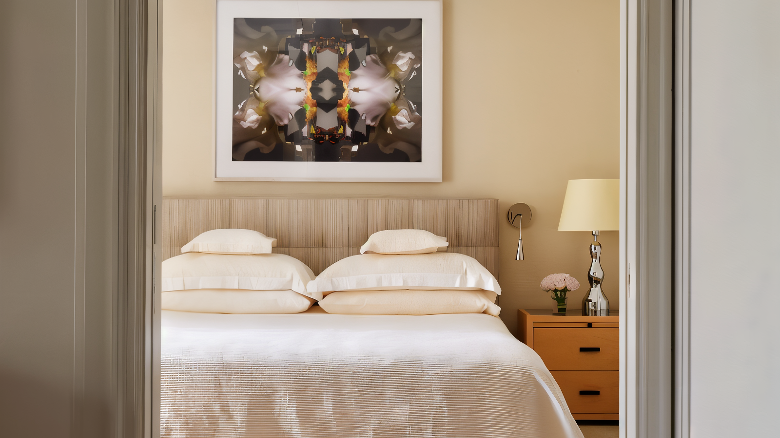 Elegant bedroom with a beige theme, featuring a double bed, abstract artwork, and a nightstand with a lamp and flowers, viewed through a doorway.
