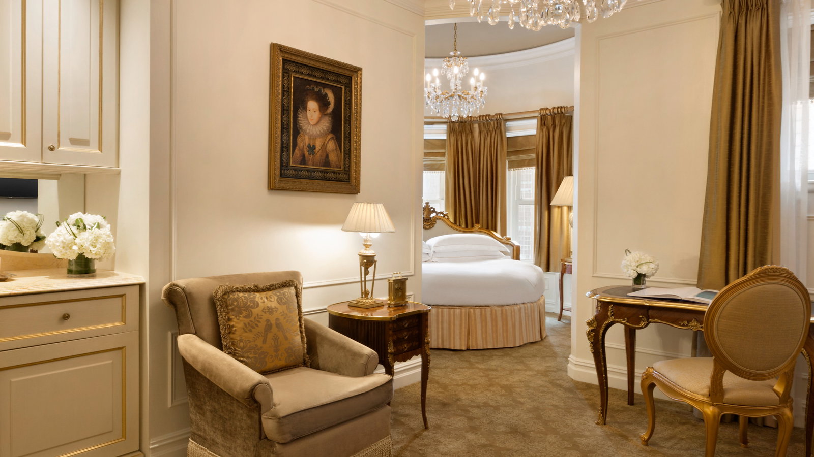 Elegant hotel room with chandeliers, golden drapery, and a framed portrait. An upholstered chair and classical desk enhance the luxurious vibe.