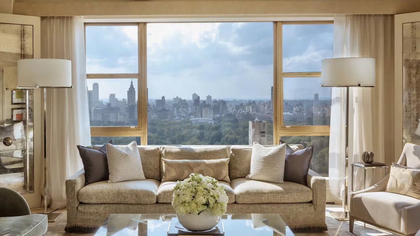 Elegant living room with beige sofa and pillows, glass coffee table with flowers, floor lamps, large window overlooking cityscape and blue sky.