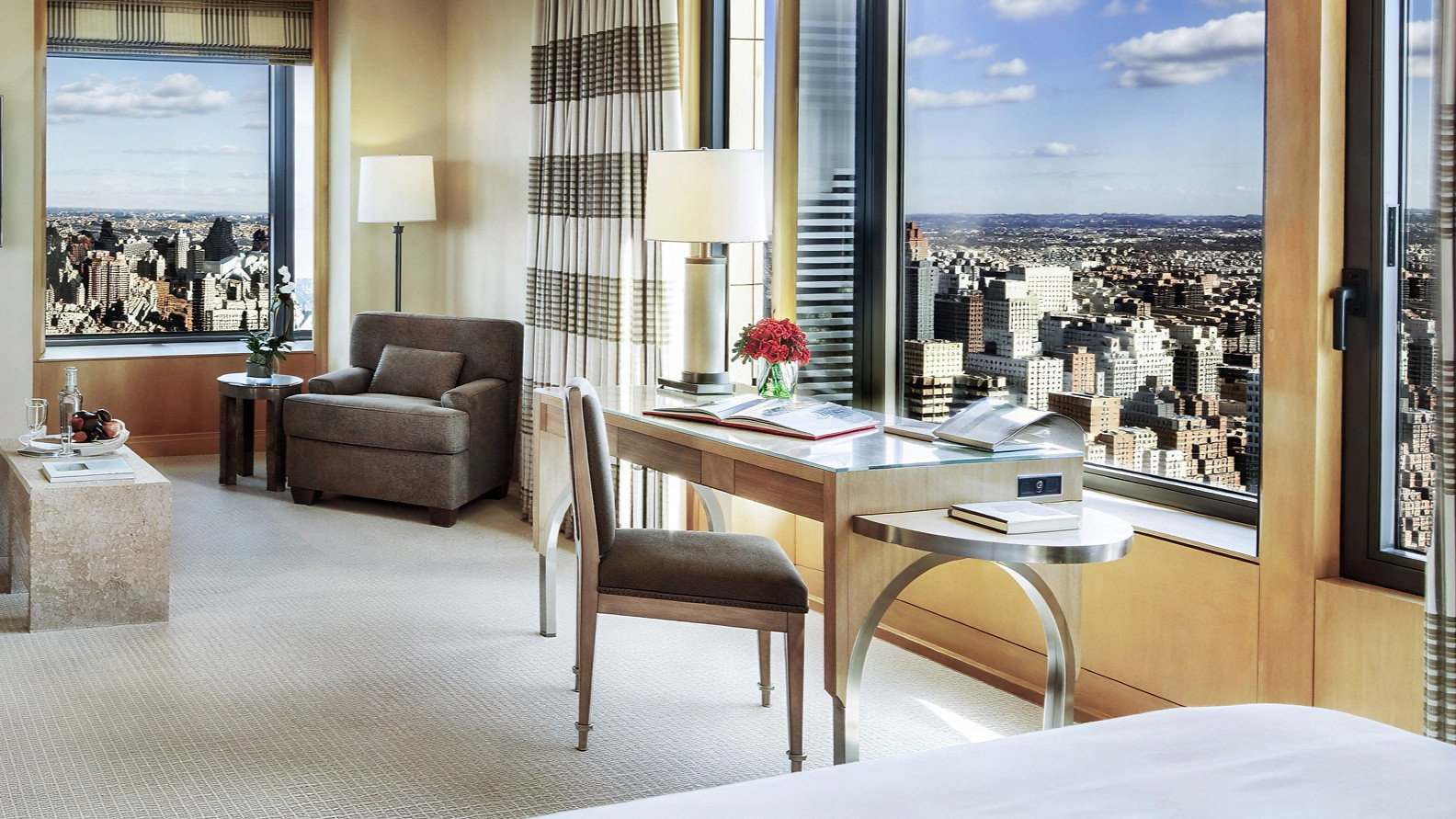 Luxury hotel room with large windows showing a cityscape. Beige tones and modern furniture, including a desk with red flowers, create a serene ambiance.