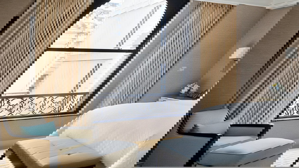 Elegant hotel room with beige armchair and ottoman, blue cushion, and white bed near window with drapes. Quiet, minimalist setting.