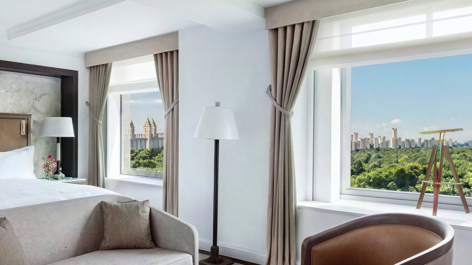 Bright, modern hotel room with neutral tones. Features large windows with city and park view, a bed, sofa, table with books, and floor lamp.