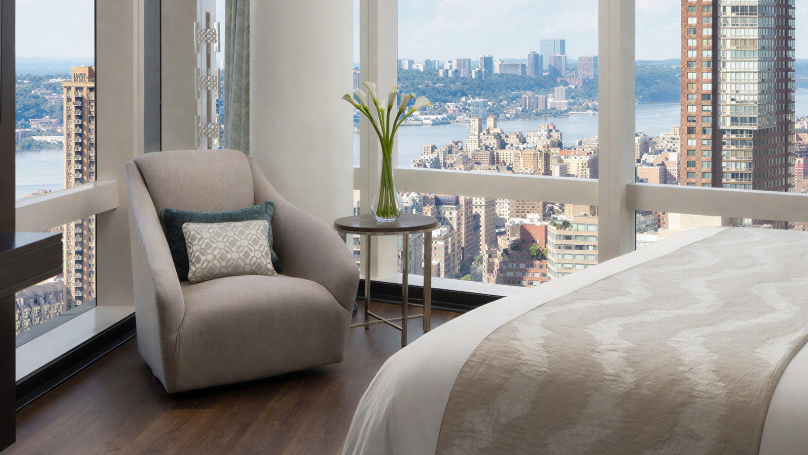 Modern bedroom with a beige armchair, patterned cushions, and a cityscape view through large windows. A vase with flowers sits on a table.