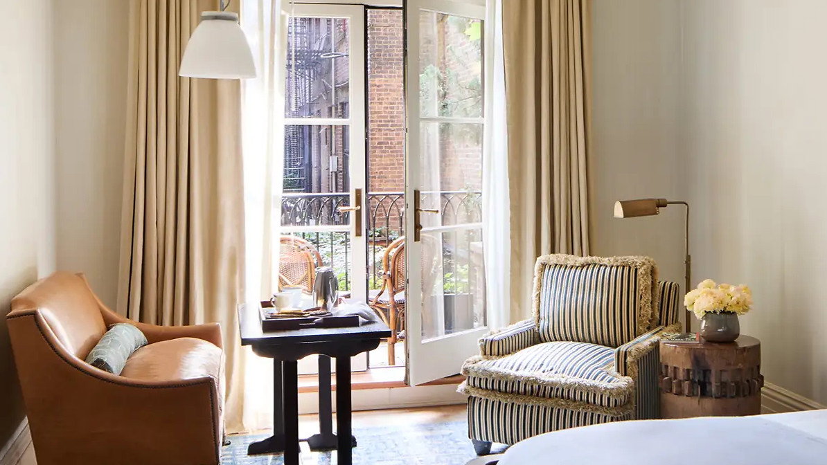 Cozy room with a brown sofa, striped armchair, and blue rug. Open French doors lead to a balcony. Neutral colors create a serene mood.