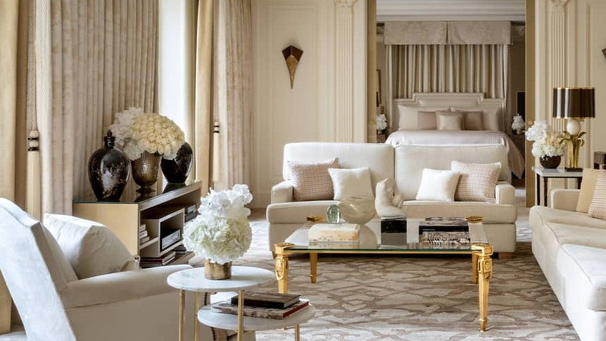 Elegant living room with beige sofas, gold-accented tables, and floral décor. Neutral tones, patterned carpet, and soft lighting evoke calm.