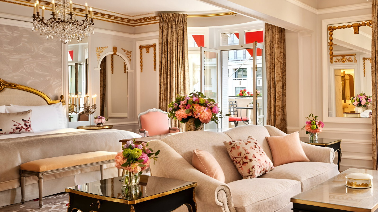Luxurious hotel suite with elegant beige furniture, floral decor, and gold accents. Sunlit balcony view, ornate mirrors, and a chandelier.