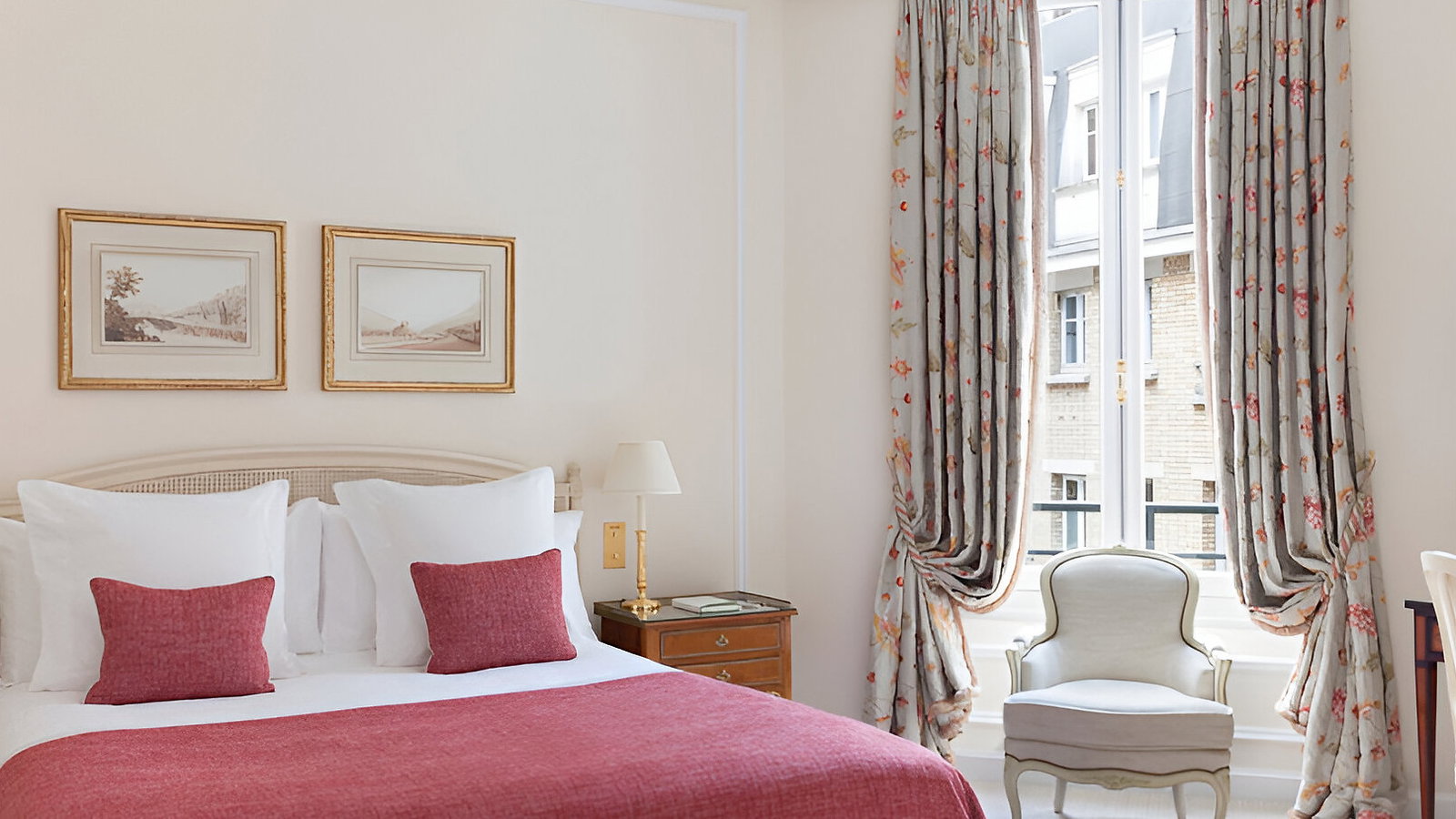 Elegant bedroom with red bedding, gold chandelier, floral curtains. Neutral walls, two framed pictures, and an upholstered chair by the window.
