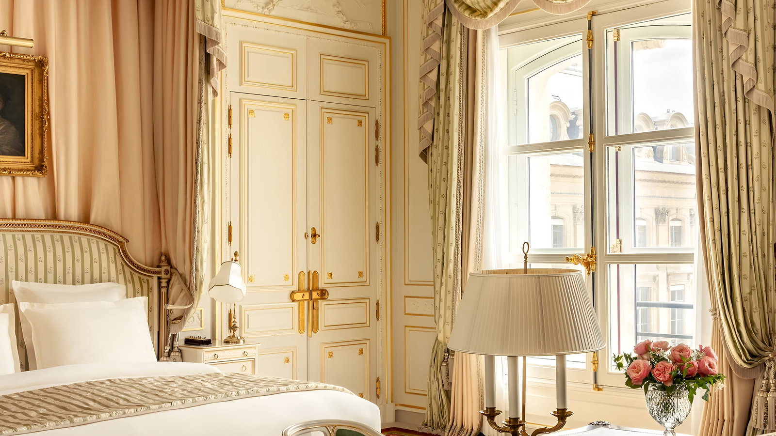 Luxurious hotel room with ornate bed, chandelier, and elegant curtains. A vase of pink roses is on a wooden table near a large window.