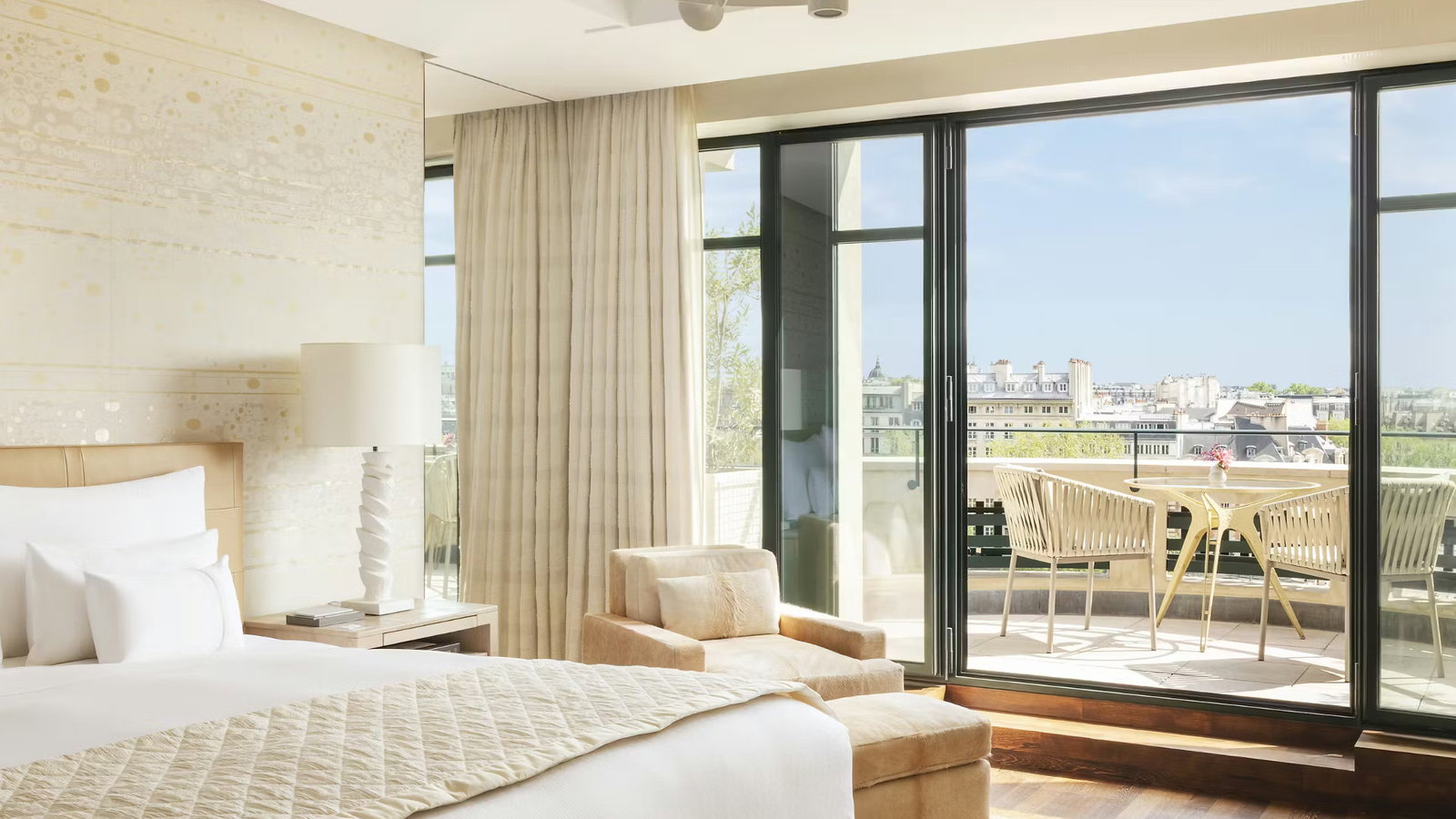 Elegant hotel room with a beige bed, armchair, and modern lamp. Sunlit balcony with a table and chairs. Large windows overlook a cityscape.