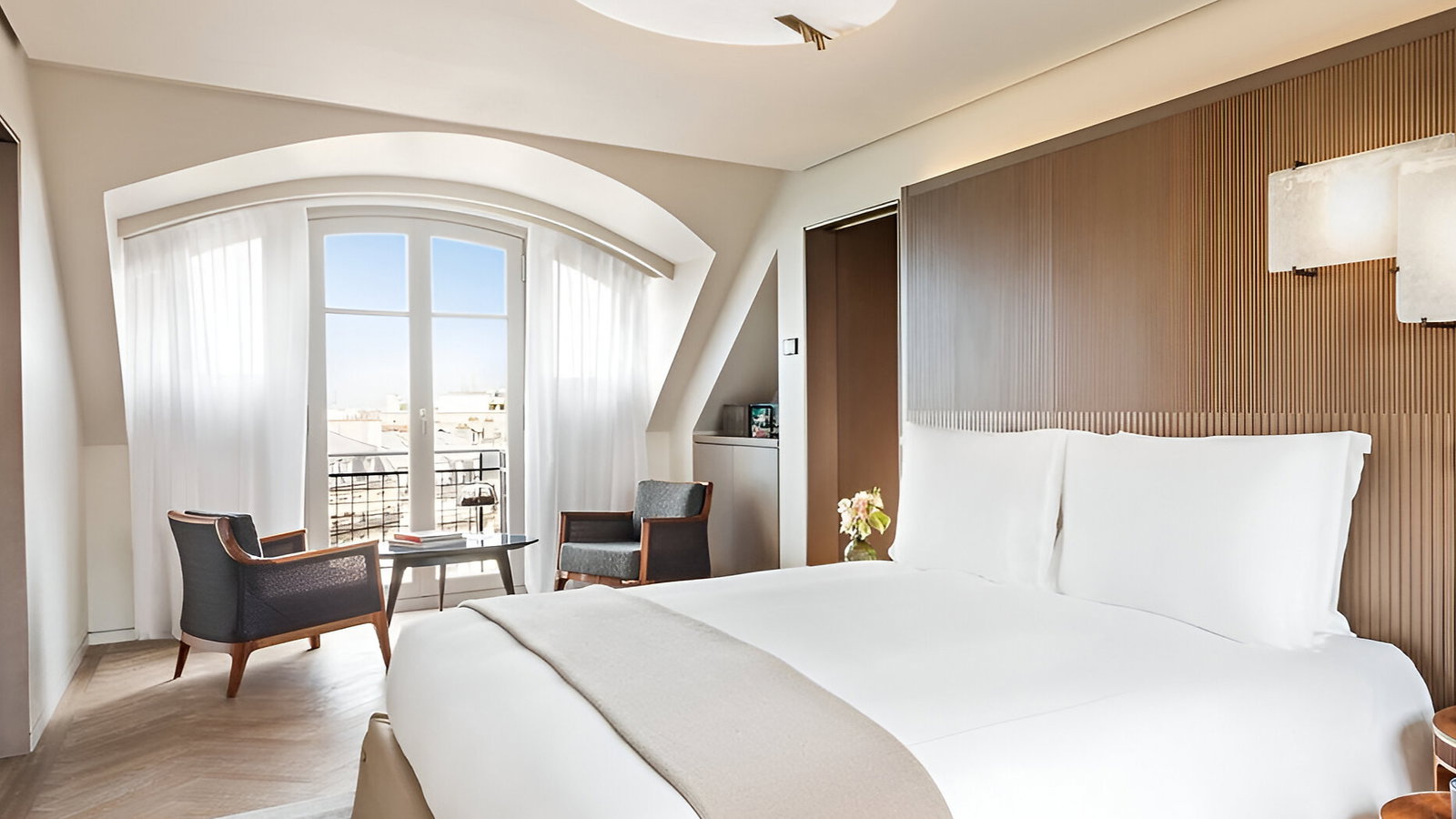 Elegant hotel room with a large bed, beige accents, two chairs by a window with sheer curtains. Soft natural light and modern decor.