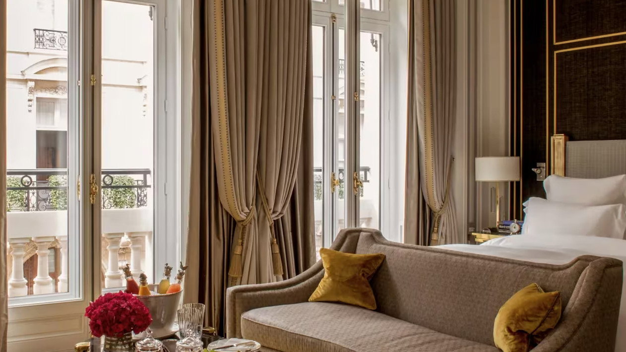 Elegant hotel room with beige sofa, gold pillows, and a glass table with fruit. Large windows with drapes and a peaceful, luxurious vibe.