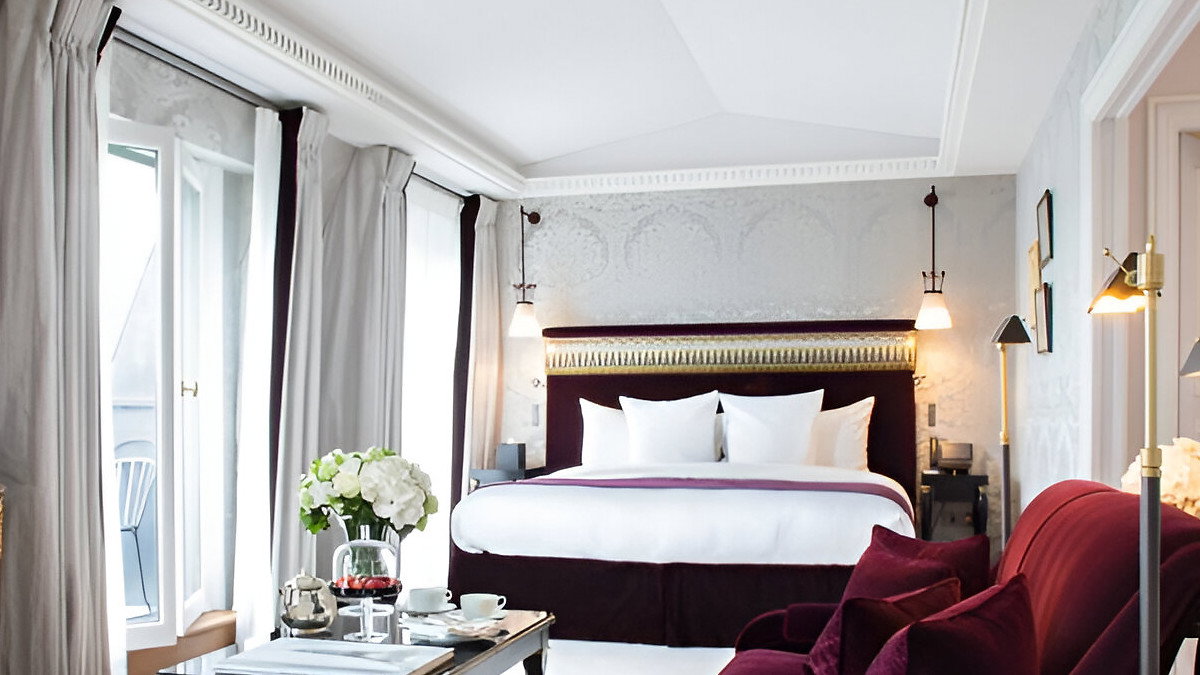 Elegant hotel room with a plush bed, maroon sofa, and a black coffee table set with tea. Soft lighting creates a cozy atmosphere.
