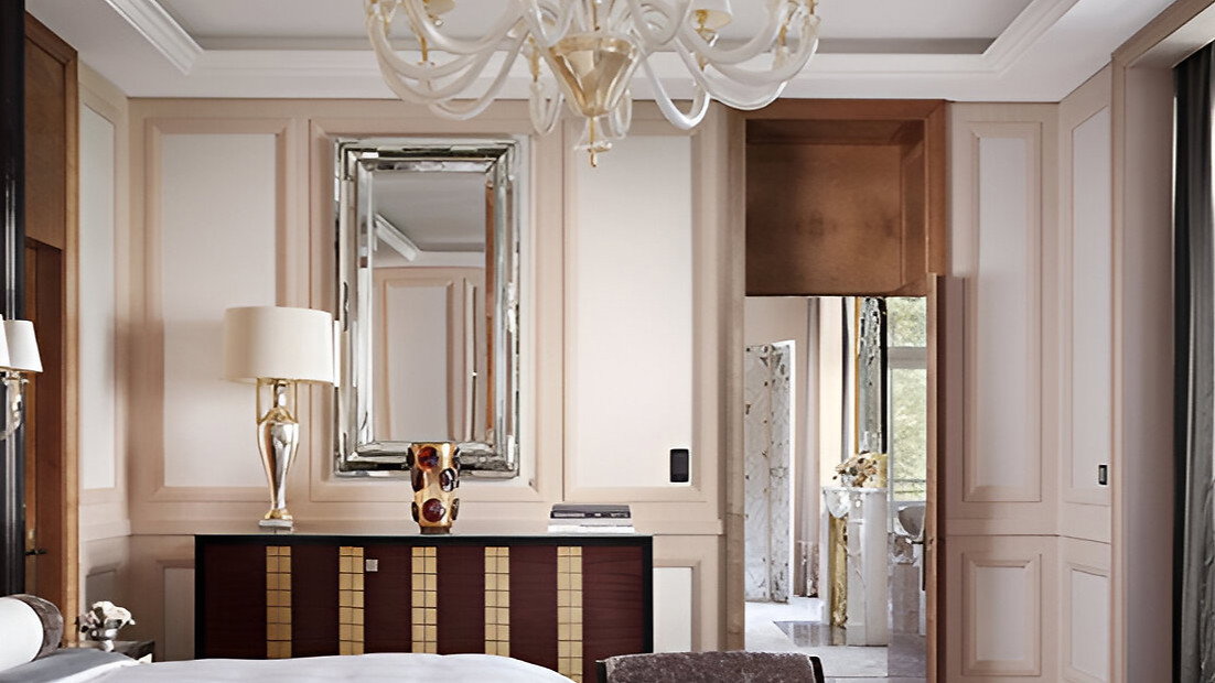 Elegant bedroom with chandelier, mirrored dresser, and plush chairs. Neutral tones dominate, with a window view of greenery. Luxurious feel.