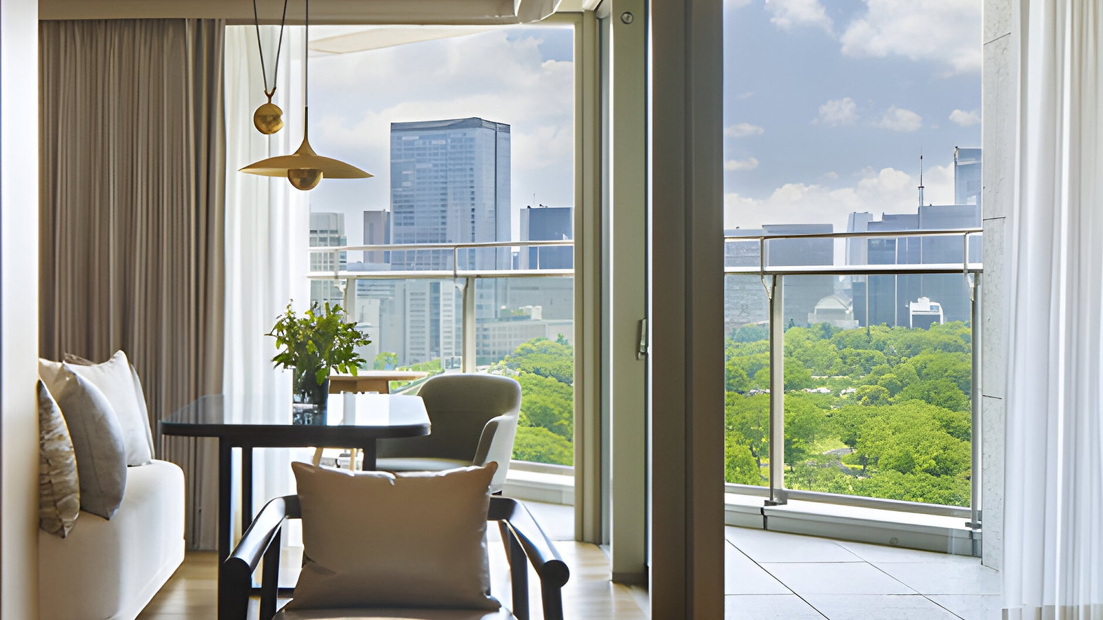 Modern living room with round marble table, sofa, and a view of city skyline through large windows. Bright, open, and stylish ambiance.