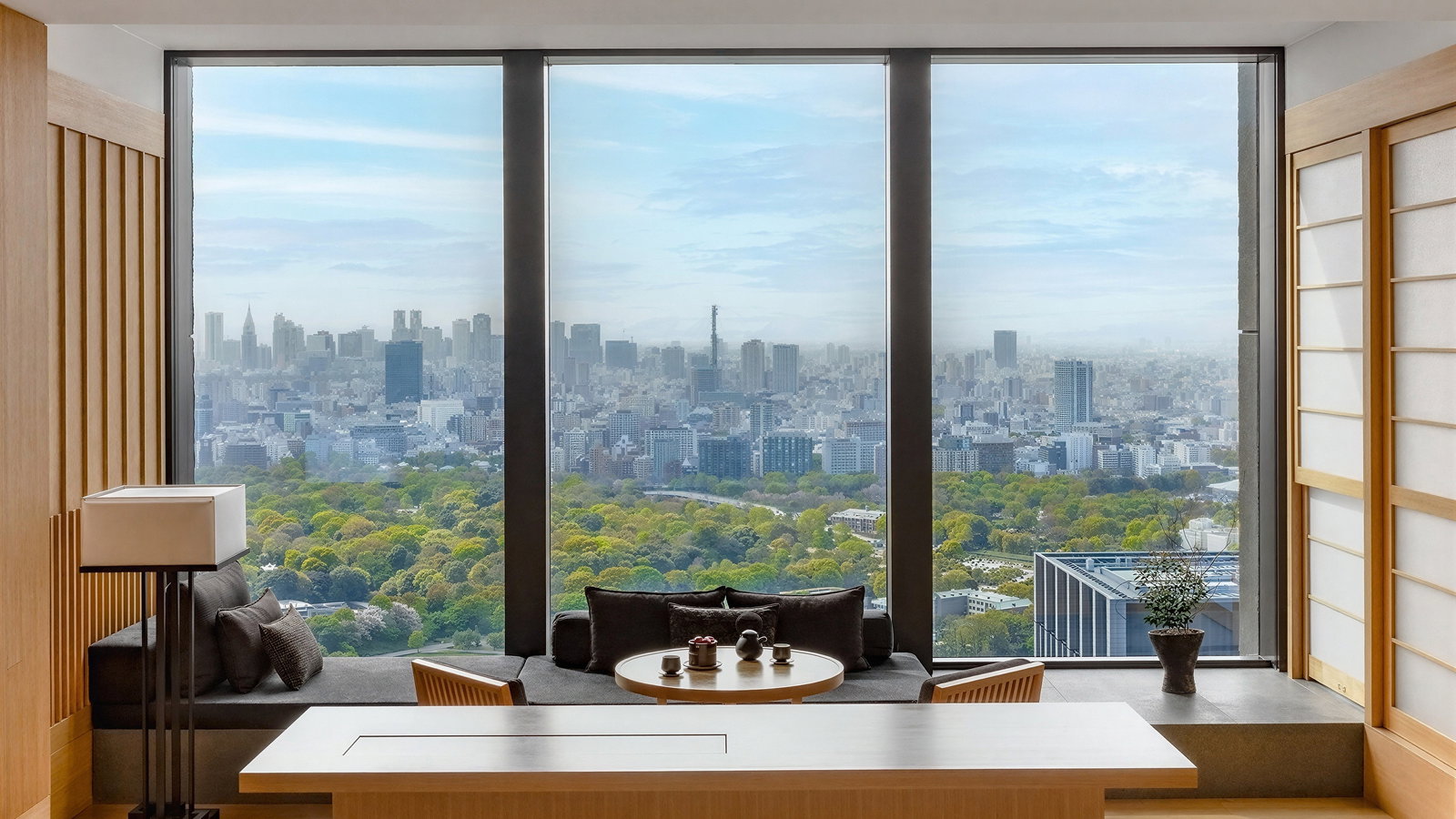 Modern room with large windows overlooking a cityscape and park. Minimalist wooden furniture and decor create a peaceful atmosphere.