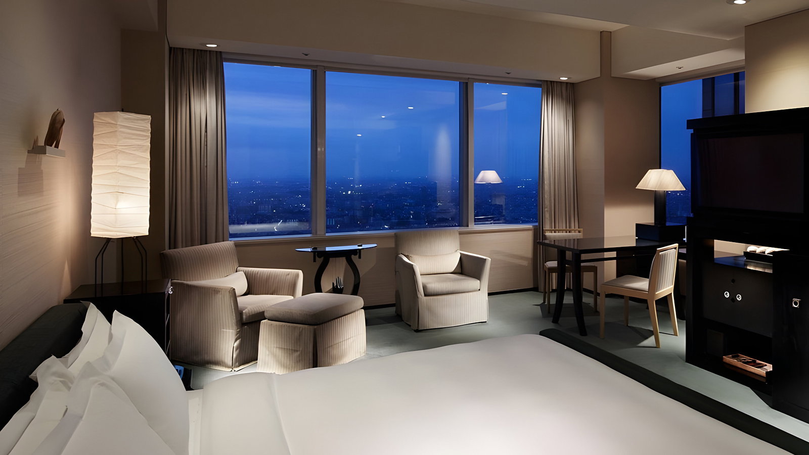 Elegant hotel room with a large bed, armchairs, and a city view through a window. Warm lighting from lamps creates a cozy atmosphere.