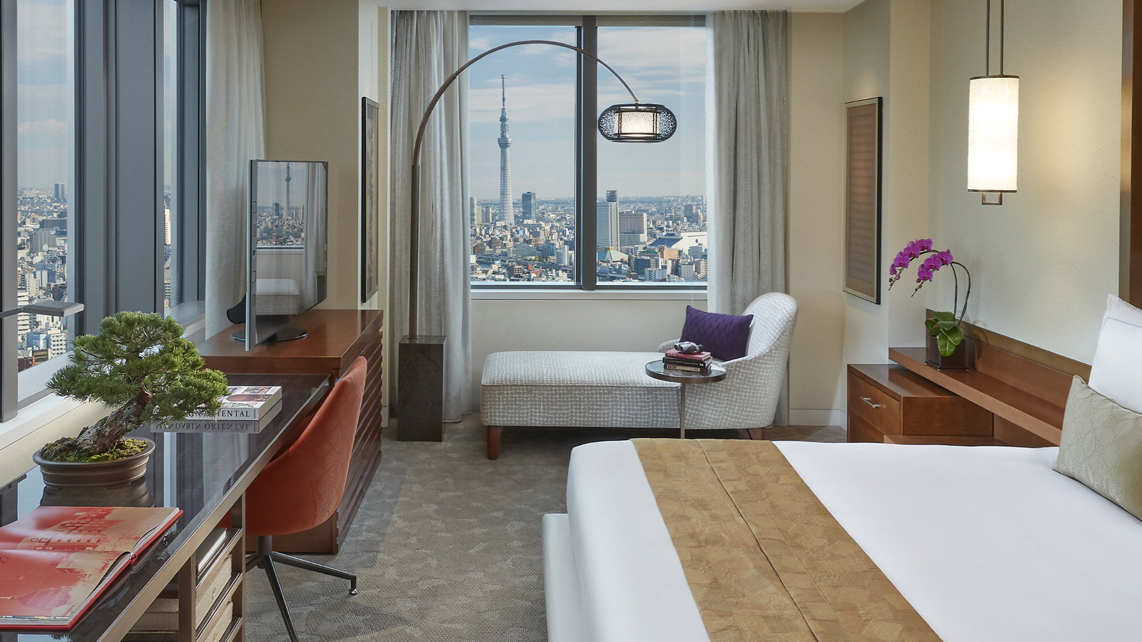 Modern hotel room with a large bed, desk, and chaise lounge. A bonsai on the desk and cityscape view through the window. Bright, elegant decor.