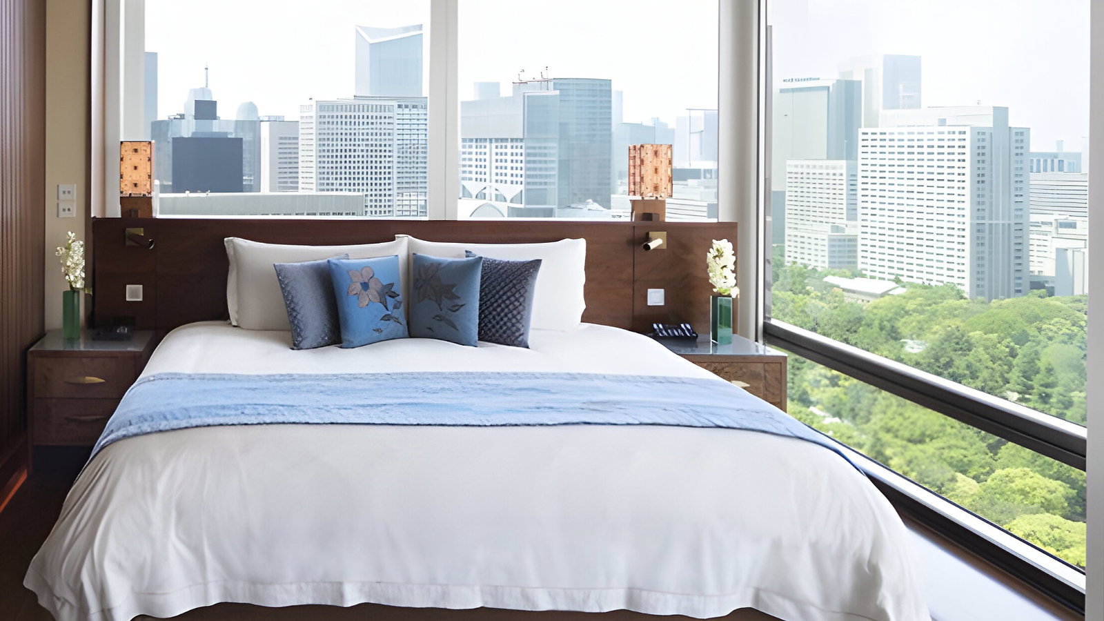 Modern bedroom with a large bed, blue pillows, and city view through large windows. Warm lighting and a blue patterned rug add elegance.
