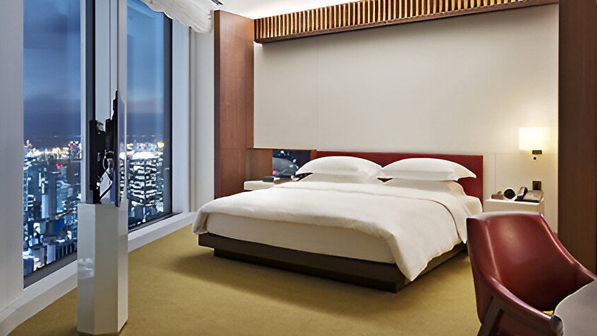 Modern hotel room with a large bed, red chair, and city view through floor-to-ceiling windows. Soft lighting creates a cozy ambiance.