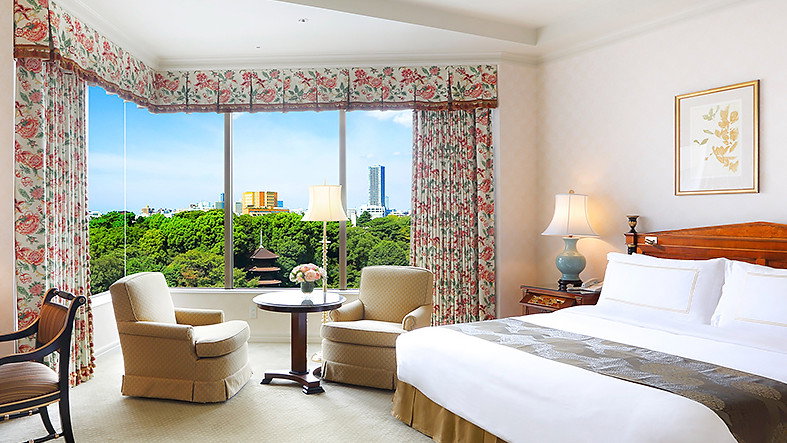 Elegant hotel room with floral curtains, two armchairs by a table, and a large bed. Window view of green trees and cityscape.