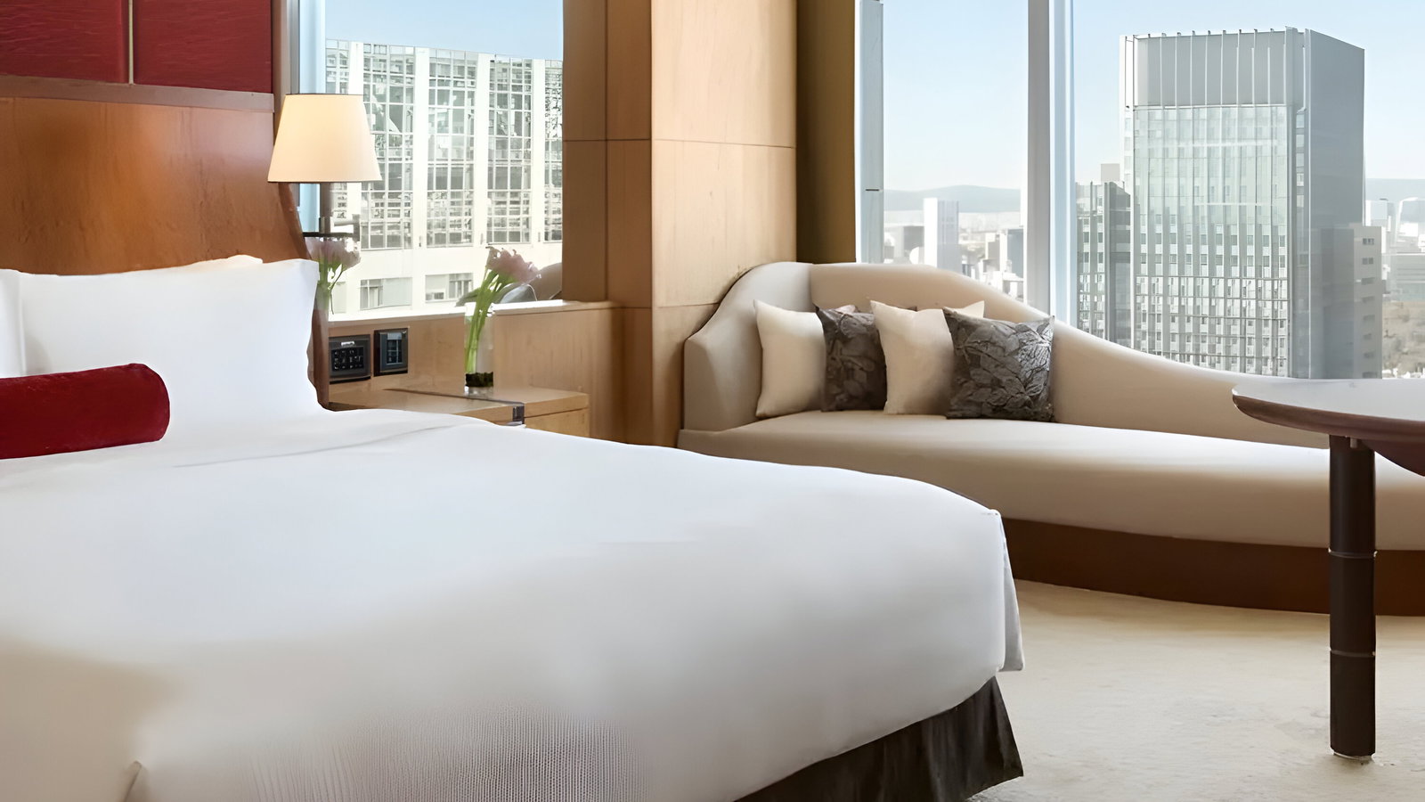 Modern hotel room with a large white bed, red cushioned headboard, beige chaise lounge, cityscape view through windows, and soft lighting.