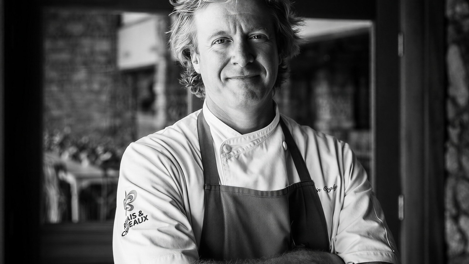 Chef in a white uniform and apron stands confidently with crossed arms. Background features a rustic interior. Black and white image.