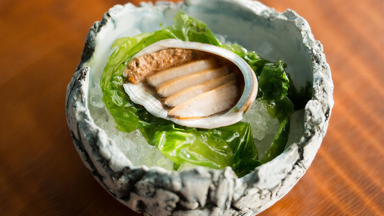 A decorative bowl on a wooden table holds a sliced mollusk on ice, garnished with green seaweed, set against a textured ceramic rim.