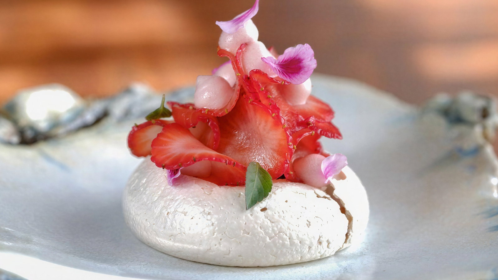 A delicate pavlova topped with sliced strawberries and pink petals sits on an elegant ceramic plate, creating a fresh and artistic dessert.