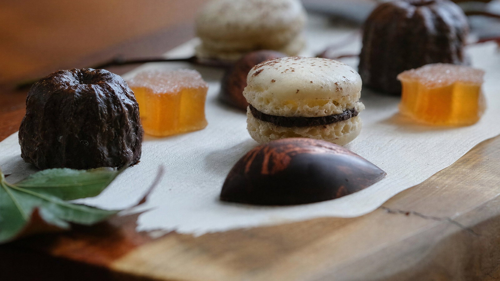 Assorted desserts on a wooden board: macarons, chocolate domes, gummy squares, and pastries. Leaves decorate the natural setting.