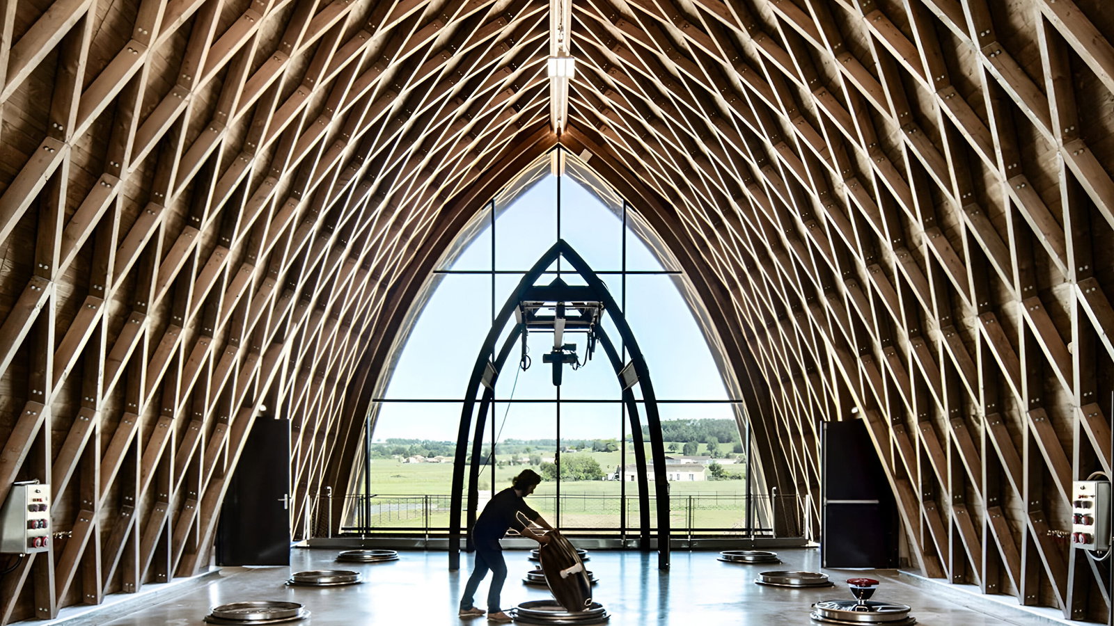 Modern wooden vaulted winery vat room interior at Château Angélus. Person moves barrel beneath a geometric roof. Bright windows reveal lush green landscape outside.