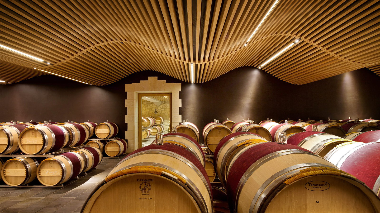 Rows of wooden wine barrels at Château Angélus in a modern barrel room cellar with a wavy wooden ceiling and soft lighting, creating a warm and elegant atmosphere.