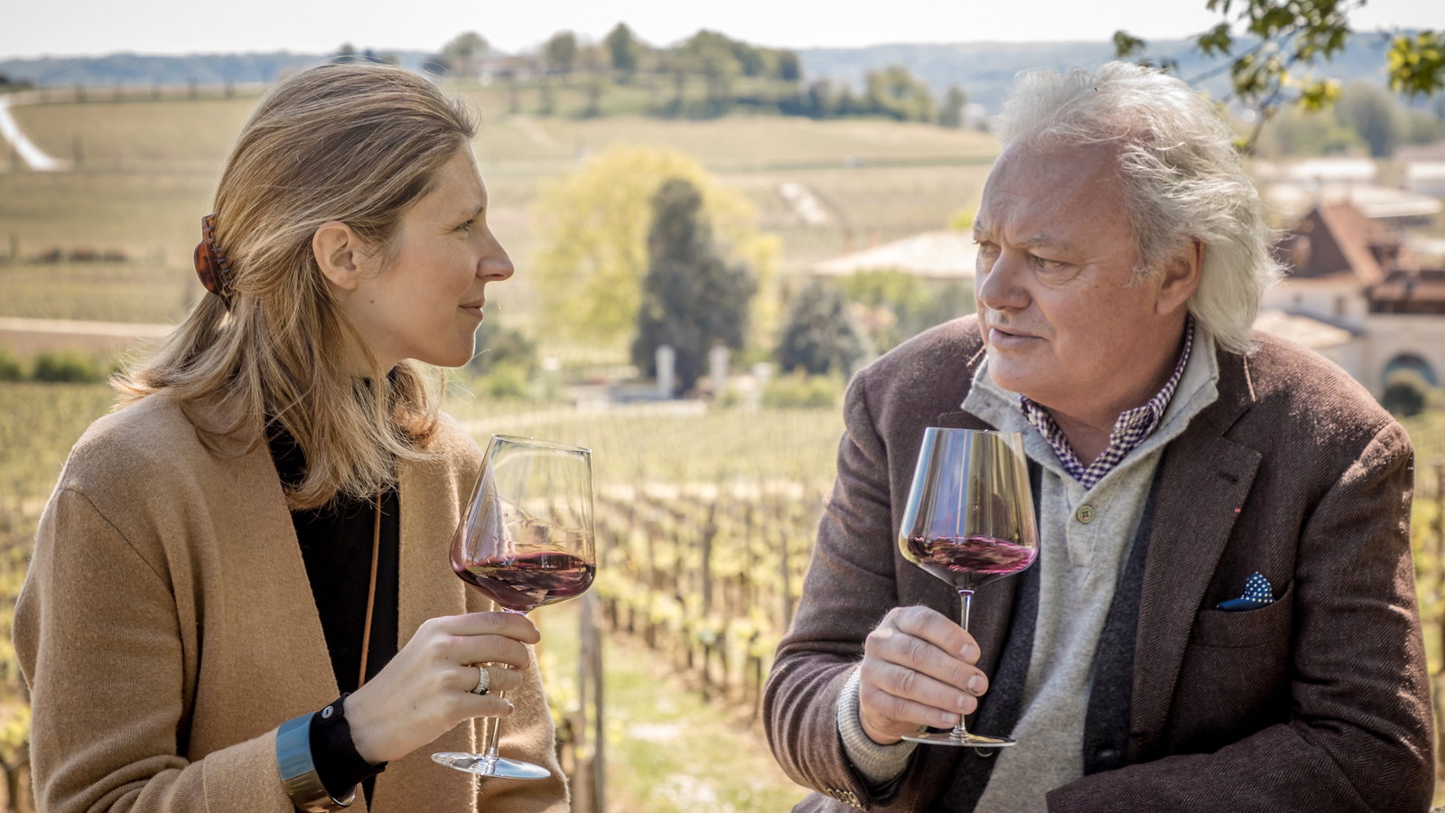Hubert de Bouard and Stephanie de Bouard hold wine glasses, conversing in the Château Angélus vineyard. Both are wearing brown coats. Rolling hills and buildings in the background.