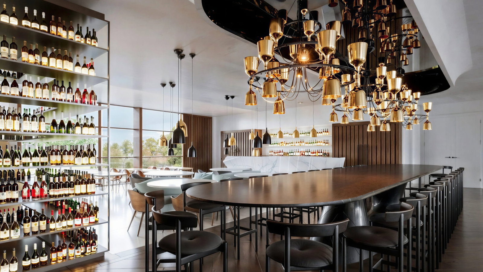 Modern wine tasting room at Carillon d'Angélus Winery, sister winery of Château Angélus with a long dark wood table, high stools, and gold chandeliers. Shelves with wine bottles line the wall, creating an elegant atmosphere.