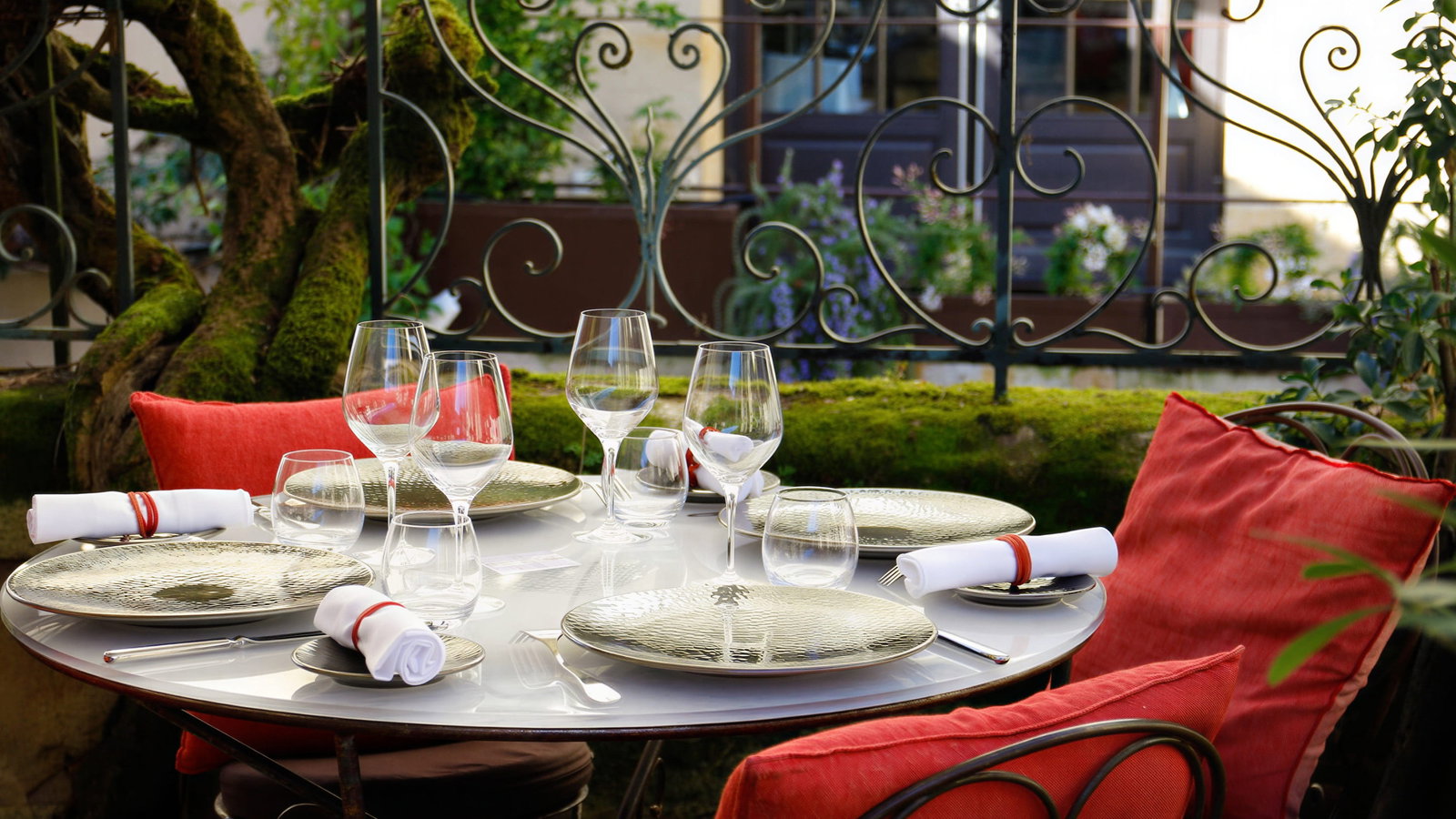 Outdoor dining table at Logis de la Cadène with empty plates, wine glasses, and rolled napkins. Red cushions on chairs, iron fence, and mossy background.