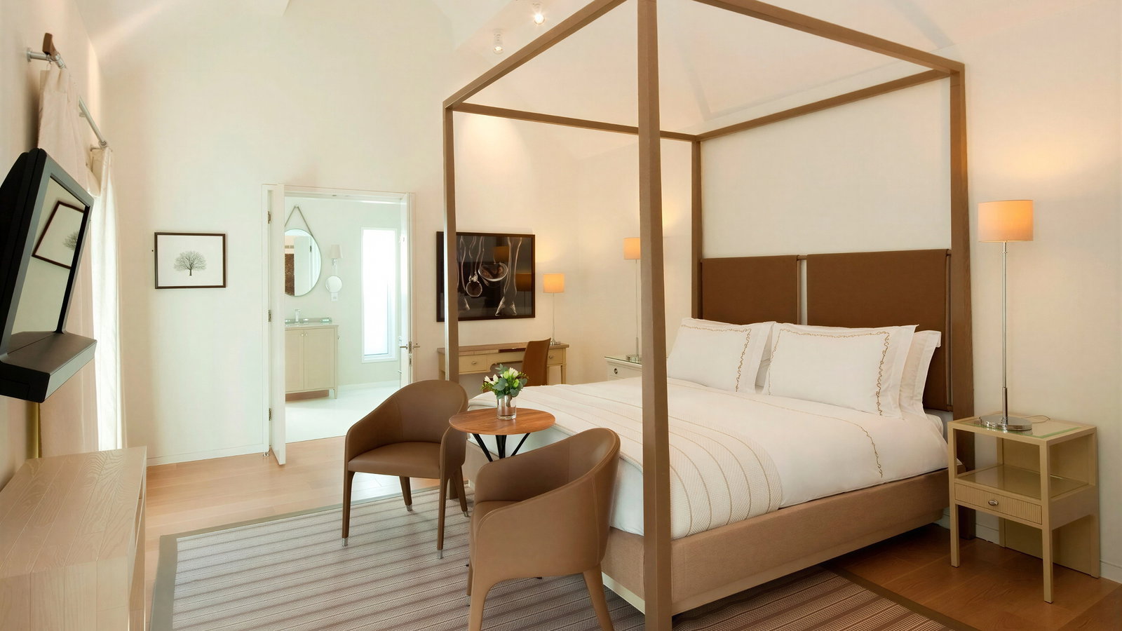 Modern bedroom with a four-poster bed, white linens, and two brown chairs around a table. Neutral tones, bright lighting, and a serene mood.