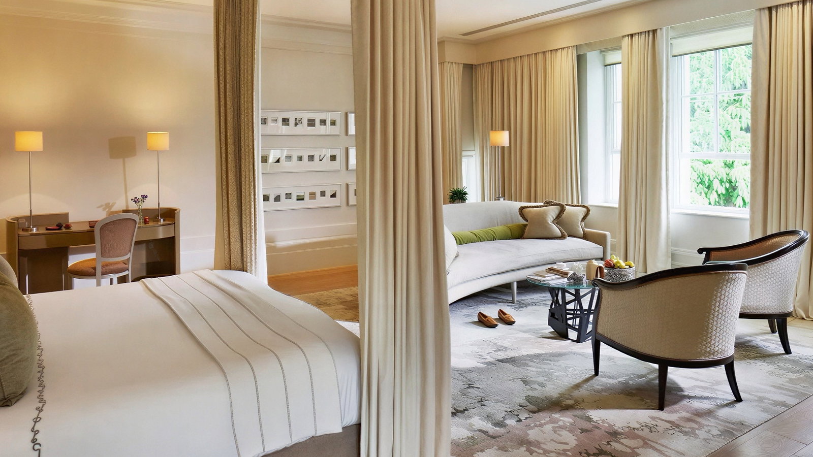 Elegant hotel room with a bed, desk, and cozy seating area. Soft beige tones, large windows, and a calm, inviting atmosphere.