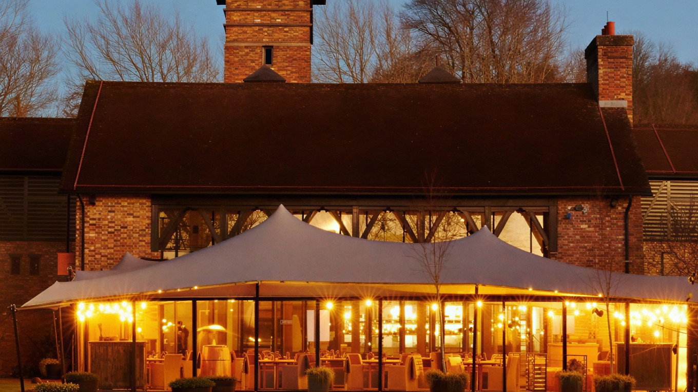 Brick building with a tall chimney at dusk, featuring a lit outdoor dining area under a canopy. Warm lights create a cozy atmosphere.