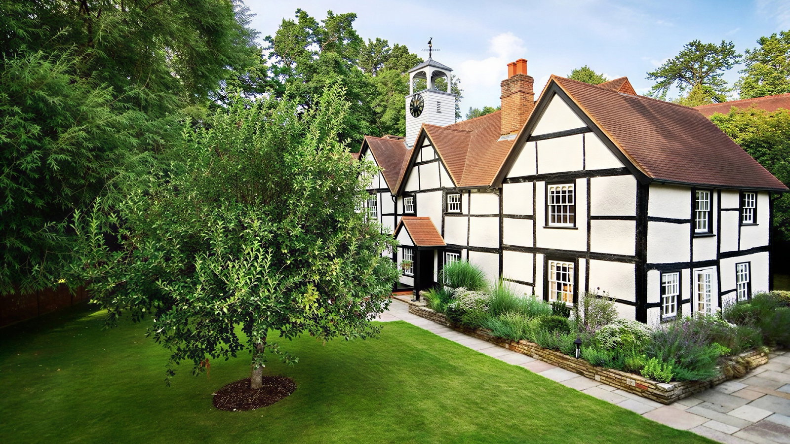 Half-timbered house with red roof, white walls, and black beams. Green lawn, lush tree, and blue sky create a peaceful setting.