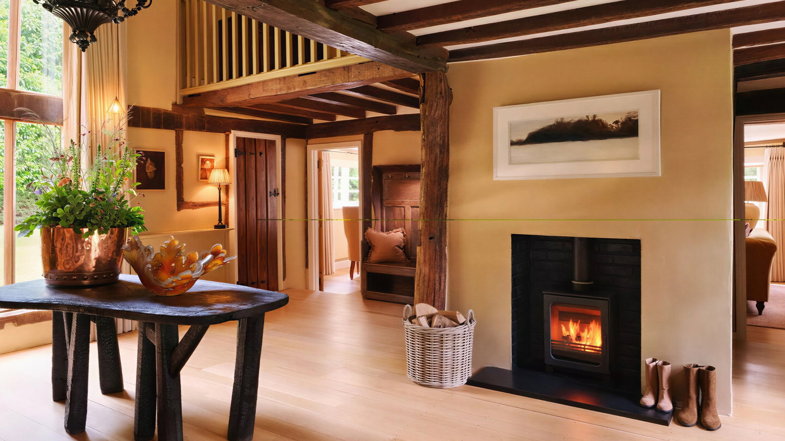 Cozy room with wooden beams, a lit fireplace, boots, and a basket. Table with copper pot and plant; warm decor with landscape art.