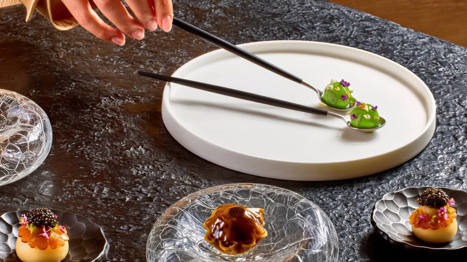 A hand places two ornate spoons of green food on a white plate. Elegant dishes with garnished food sit on a textured black table.