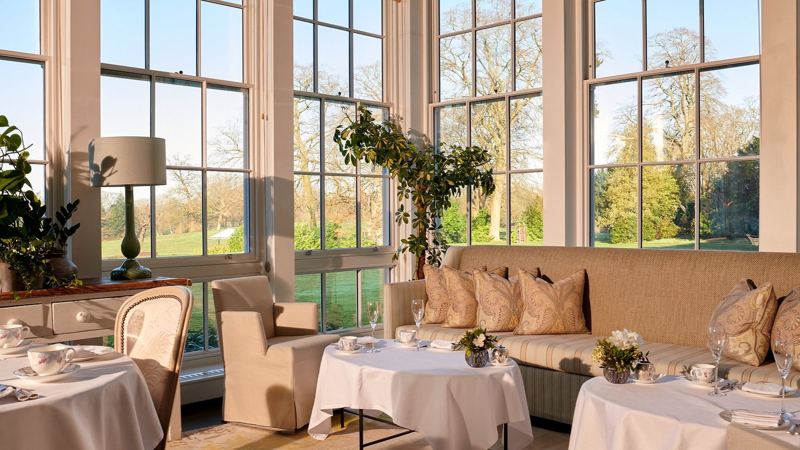 Sunlit dining room with large windows, a beige sofa with patterned pillows, white tablecloths, and green garden view. Calm and inviting.